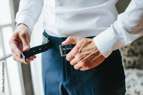  A young man fastens a black belt on his trousers, male hands close-up. Morning of the groom, preparations for the wedding. Selective focus.