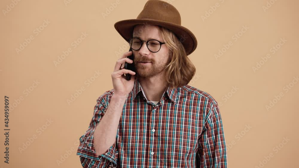A confident man in the hat and eyeglasses talking on the phone and shaking his head negatively while standing in a beige studio A confident man in the hat and eyeglasses talking on the phone and shaking his head negatively while standing in a beige studio