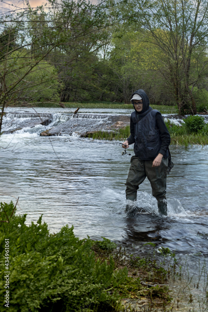 fisherman in vest, cap and hooded sweatshirt wading in the river with a fishing rod and tall rubber boots as he emerges from the stream
