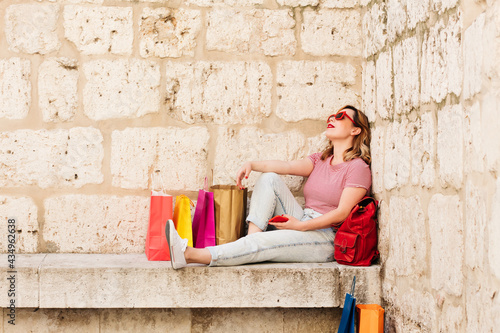 Woman with red sunglasses resting in the street of her city surrounded by colorful bags concept addicted to shopping, sales and offers. Lifestyle.