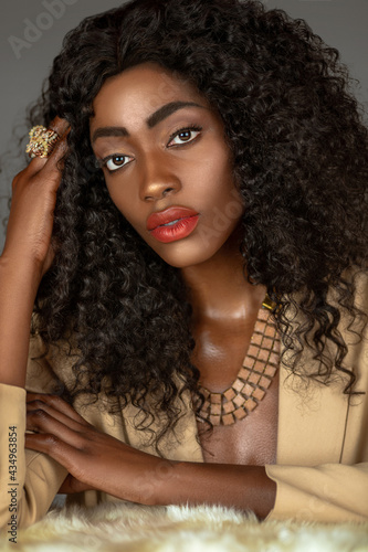 Closeup portrait of a beautiful black woman with long curly black hair and beautiful makeup posing by herself on a fur in a studio with gray background wearing a beige suit and jewelry.