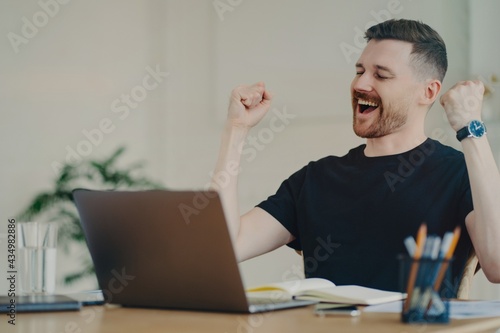 Happy young freelancer showing winning gesture while sitting at his workplace at home photo