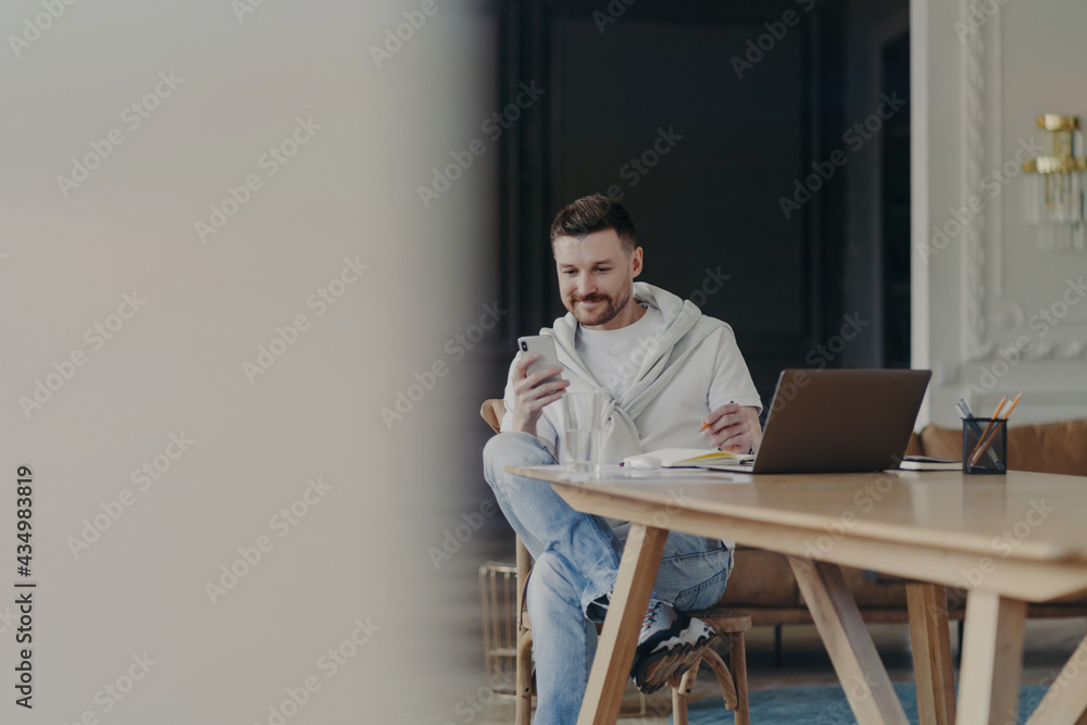 Joyful young male entrepreneur sitting near wooden table and using smartphone while working remotely on laptop from home Joyful young male entrepreneur sitting near wooden table and using smartphone while working remotely on laptop from home