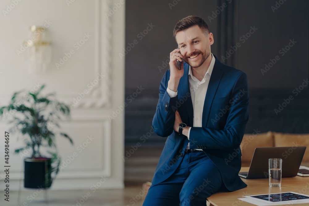 Smiling successful CEO talking on phone while sitting on table in office Smiling successful CEO talking on phone while sitting on table in office