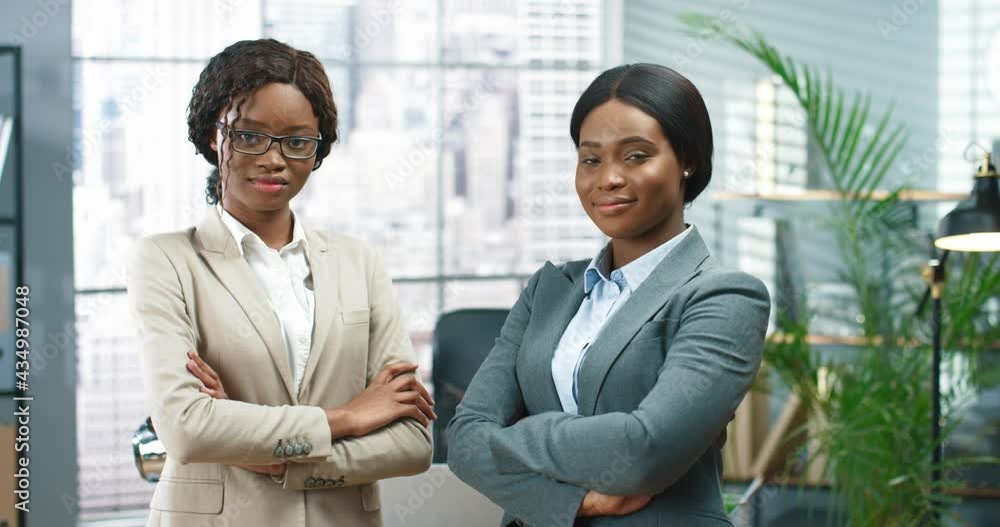 Portrait of African American young beautiful female employees in suits standing in office room and looking at camera. Happy businesswomen at work in cabinet, colleagues work in company