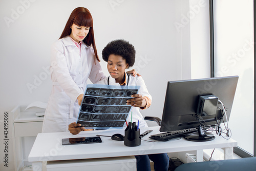Two young multiethnic women, African and Caucasian, professional doctors oncologists radilogists, look and discuss an X-ray or MRI scan of the patient, working in light modern clinic