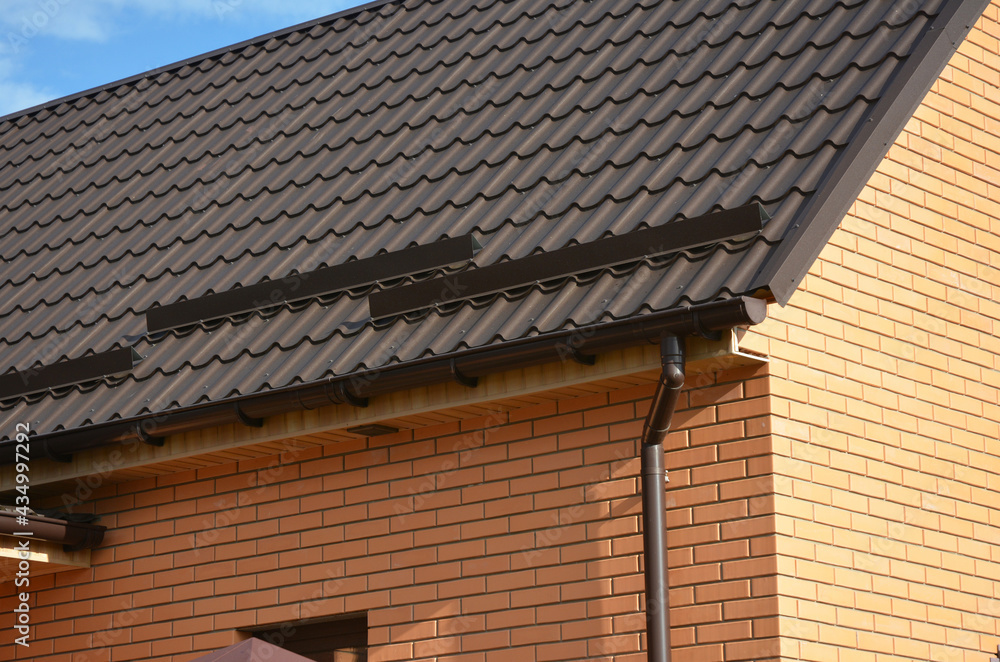 A close-up on a metal brown roof of a brick house with snow stoppers, snow guards, a rain gutter, a downspout, soffit and fascia board.
