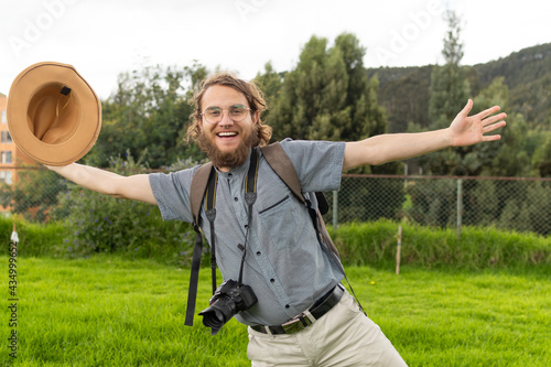 young traveler with glasses posing amused smiling carrying his camera and holding his hat with outstretched arms