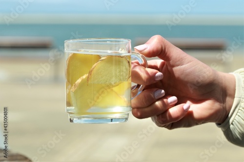 Ginger tea with slices of ginger hold by a hand in front of a beach