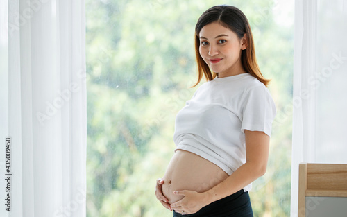 Free Download portrait of a good-looking Asian pregnant woman dress casually standing and touching her belly with love and happiness while looking and smiling at a camera - GFXLoad