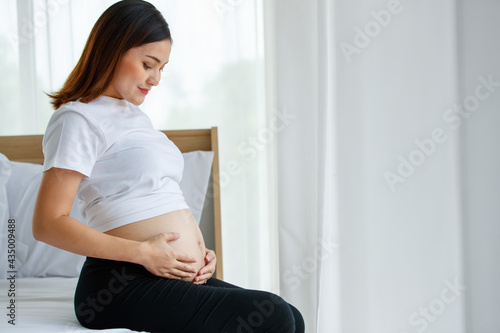 happy smiling young beautiful Asian pregnant woman sitting on a bed looking and touching her belly with care and love. Expecting for a strong and healthy baby