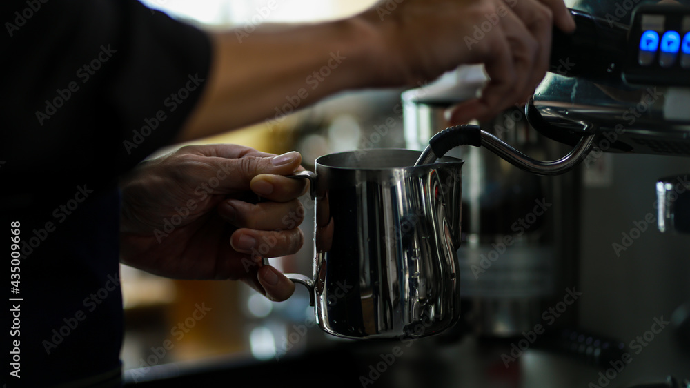 Close-up shot of stainless cup holding by two hands of barista while pouring hot water from steel luxury coffee making machine with blurred cafe background