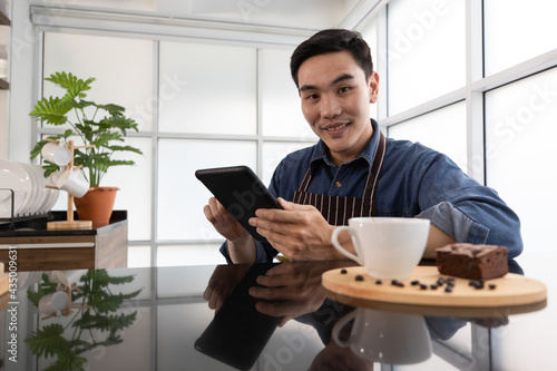 Close up shot of Asia short black hair handsome barista wearing longsleeve blue jeans shirt with brown stripe apron sitting smiling and using black tablet at coffee shop table with coffee and brownie