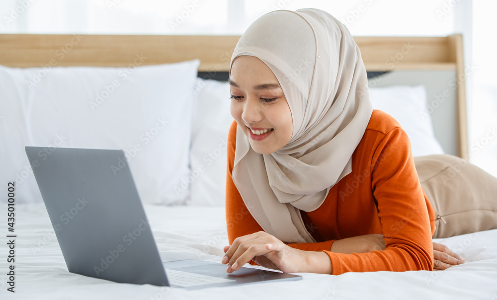 A cute and beautiful Asian Muslim in hijab laying on bed and using tablet notebook computer A cute and beautiful Asian Muslim in hijab laying on bed and using tablet notebook computer