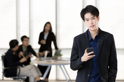 An executive look tomboy woman in casual suit standing with a self-confident and using smartphone in office and colleagues in background. Transgender in modern business workplace concept
