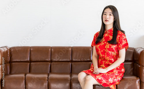 Chinese beautiful woman with long black hairs in red traditional dress is sitting politely on a brown sofa and put hands on her lap photo