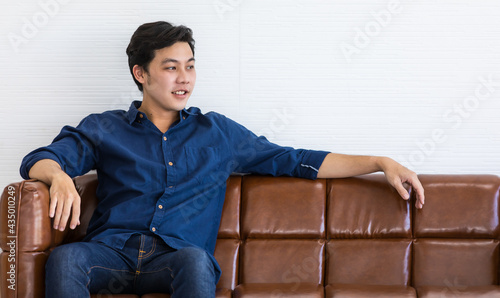 Young man in blue shirt and blue jeans is sitting on a dark brown sofa with smile. He stretch his left arm out for relax. © Bangkok Click Studio
