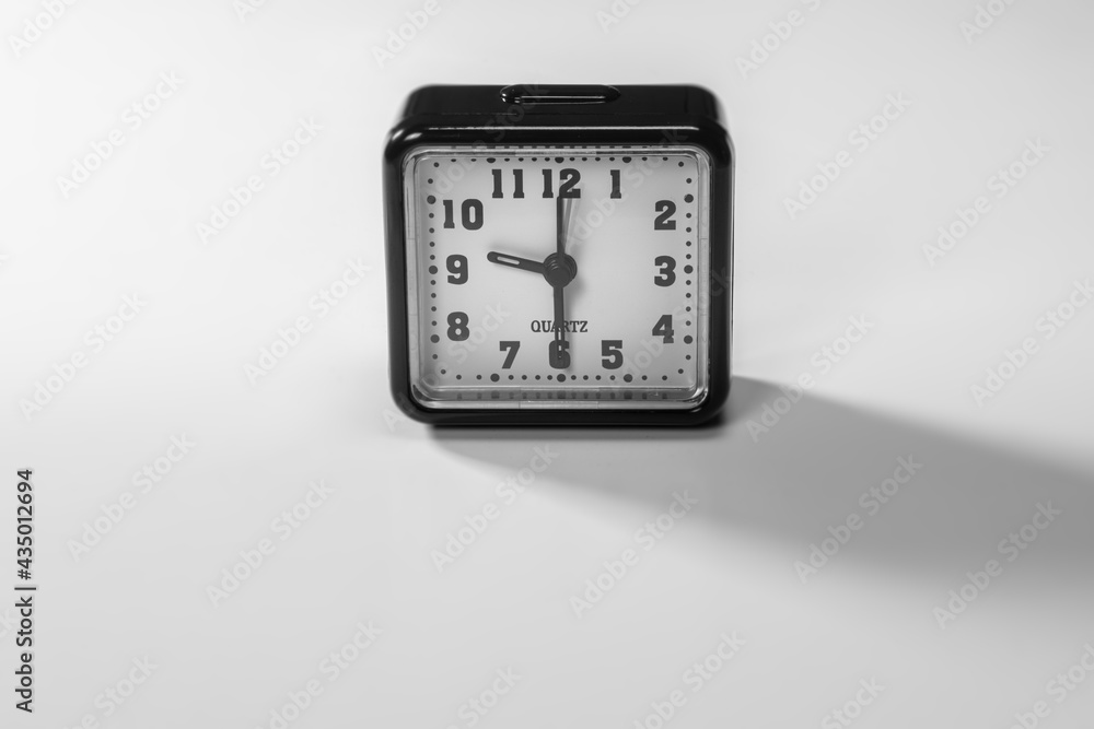 Black alarm clock on white background. The long shadow from the sun. Traditional alarm watch with black plastic frame. Long shade from the rising sunlight. Morning sun on a simple alarm chronometer Black alarm clock on white background. The long shadow from the sun. Traditional alarm watch with black plastic frame. Long shade from the rising sunlight. Morning sun on a simple alarm chronometer