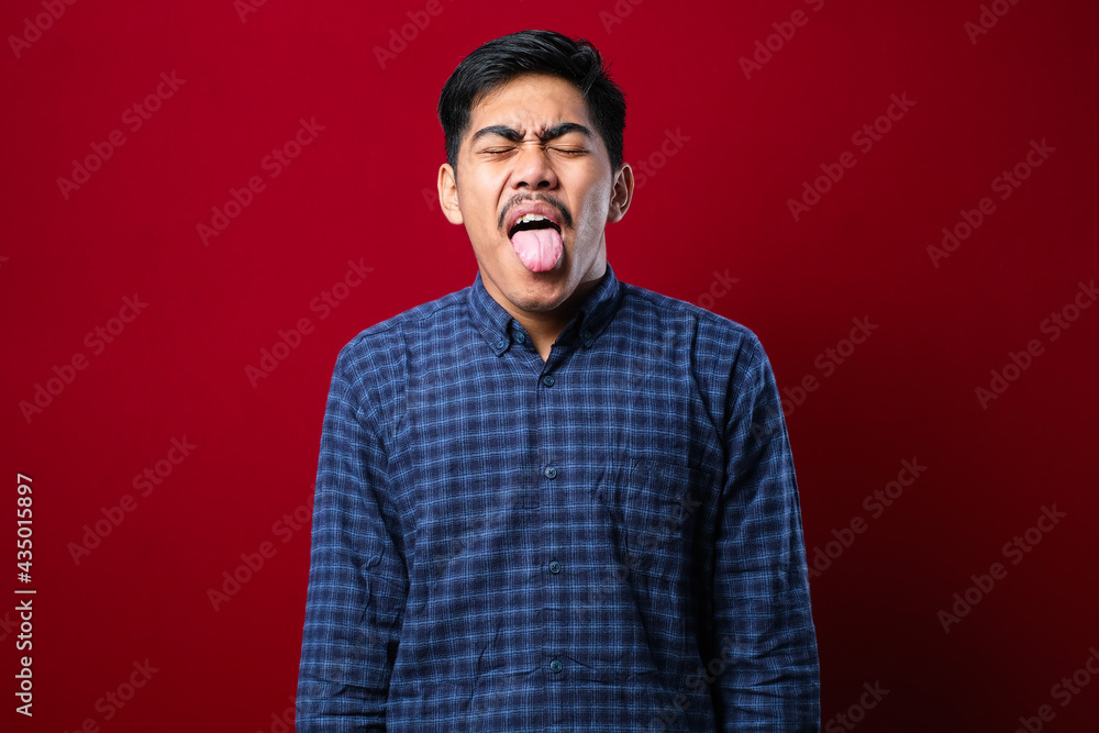 Young handsome asian boy wearing casual shirt standing over red background sticking tongue out happy with funny expression.