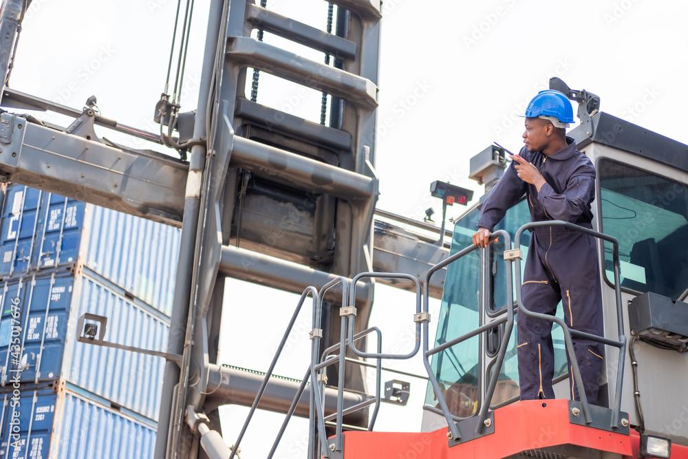 Selective focus at face of Black African logistic worker wearing safety equipment, check and inspection while loading container while talking and communicate with his team by  radio device.