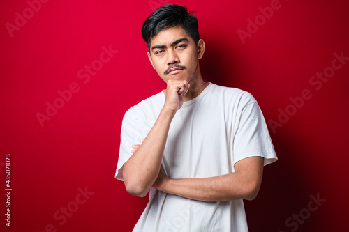 Handsome asian man with mustache wearing white tshirt looking confident at the camera with smile with crossed arms and hand raised on chin. Thinking positive.