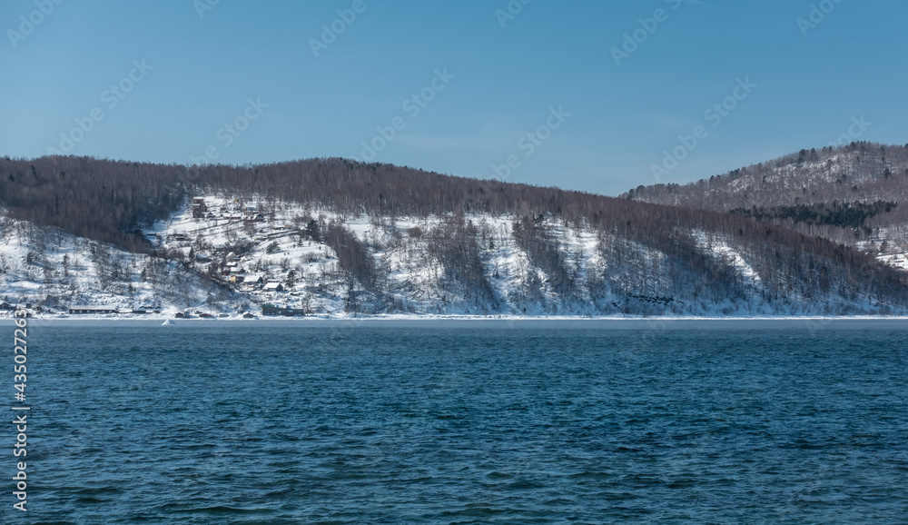 The non-freezing Angara river. Ripples on the blue water. On the shore, on the slope of a snow-covered mountain, one can see village houses and a forest. Clear sky, sunny winter day.