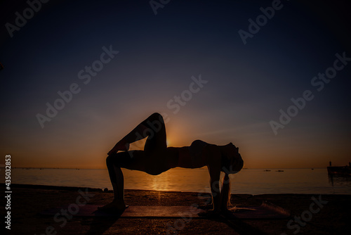 Young blonde woman in sportswear performs yoga asanas, exercises on the seashore at sunrise. The girl goes in for sports, works out breathing, meditates, performs Reverse bridge pose. Healthy mind