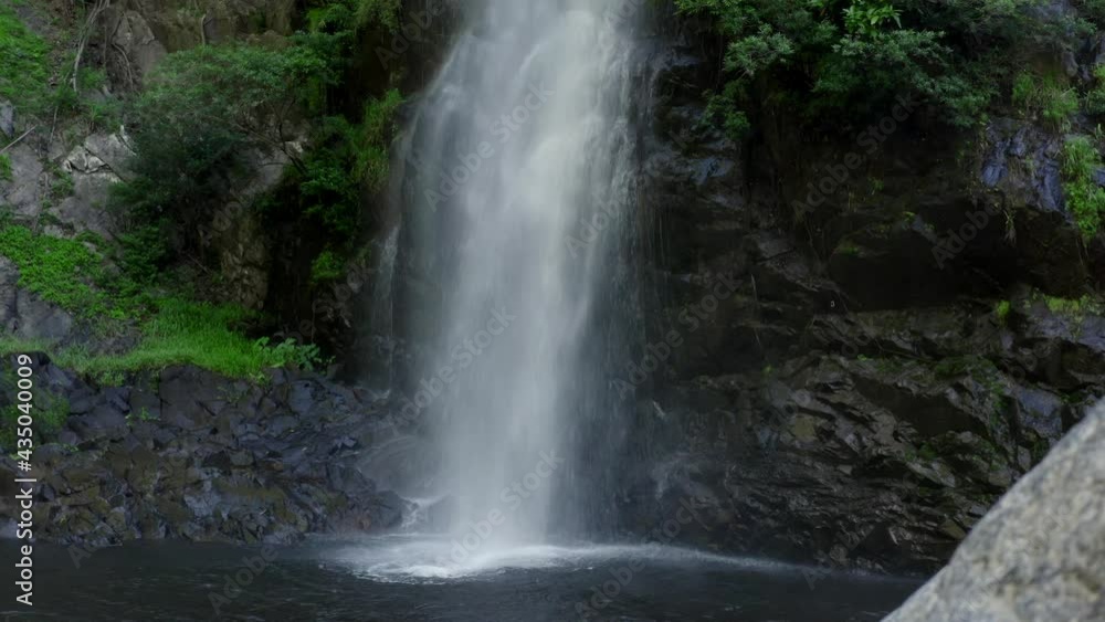 Beautiful landscape with a waterfall surrounded by green trees. The camera stands on the shore and we see how powerful jets of water fall into the lake. there is a large stone in the foreground Beautiful landscape with a waterfall surrounded by green trees. The camera stands on the shore and we see how powerful jets of water fall into the lake. there is a large stone in the foreground