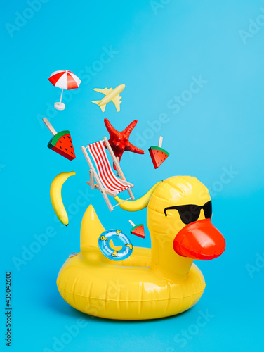 Yellow inflatable floating duck with flying beach chair, starfish, ice cream and and plane on a blue background. Creative trendy summer vacation concept. © Aleksandar