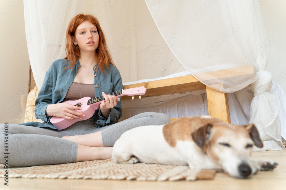 young beautiful red haired girl playing ukulele small pink guitar in bedroom. Chilling home with dog Jack russell terrier out of focus in the foreground. Blue linen shirt. Weekend mood new skills