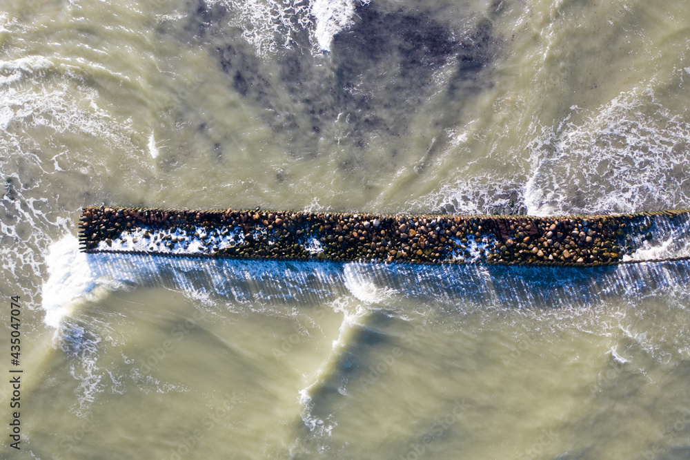 Obraz premium Aerial top down view of waves breaking against old stone breakwater pier in sea