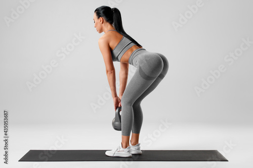 Athletic girl doing deadlift exercise for glutes on gray background. Fitness woman working out with kettlebell