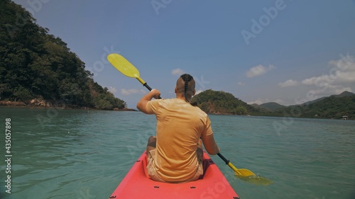 Man with sunglasses and hat rows pink plastic canoe along sea against green hilly islands with wild jungles. Traveling to tropical countries.