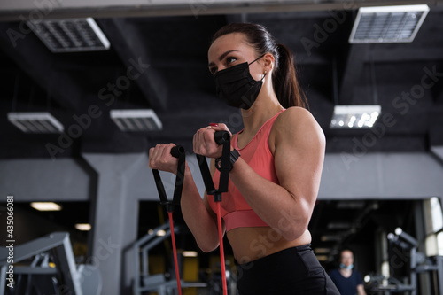 Low angle shot of a sportswoman doing resistance band biceps curls  wearing medical face mask at gym