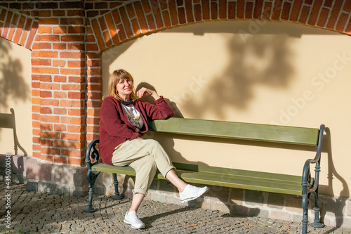 middle-aged woman, cheerful pretty sits on a bench against the background of a wall and smiles