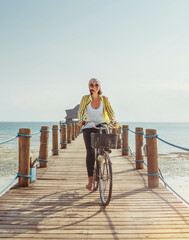 Portrait of a happy smiling woman dressed in light summer clothes and sunglasses riding a bicycle on the wooden sea pier and looking at camera. Careless vacation in tropical countries concept image