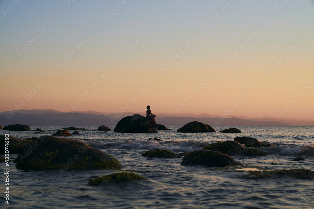 Silhouette of Peaceful woman meditating on a rock in the middle of a low wave sea at sunset time. golden hour colorr
