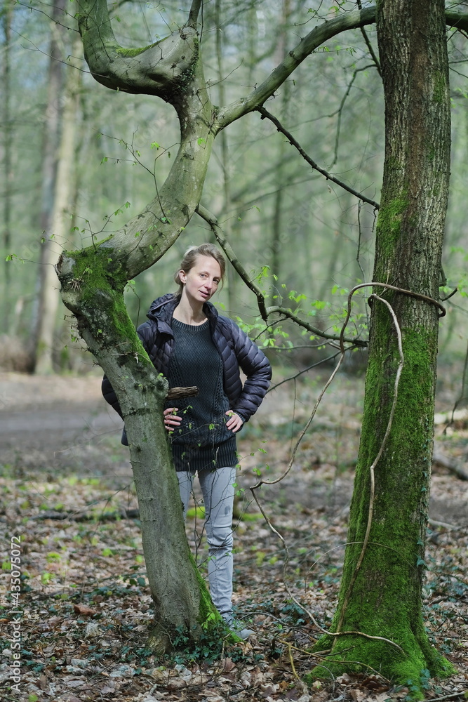 Eine junge blonde, schlanke Frau in helle jeans und dunkle blaue Jacke an einem Baum mit wenige hell grüne Blätter in Wald. Eine junge blonde, schlanke Frau in helle jeans und dunkle blaue Jacke an einem Baum mit wenige hell grüne Blätter in Wald.