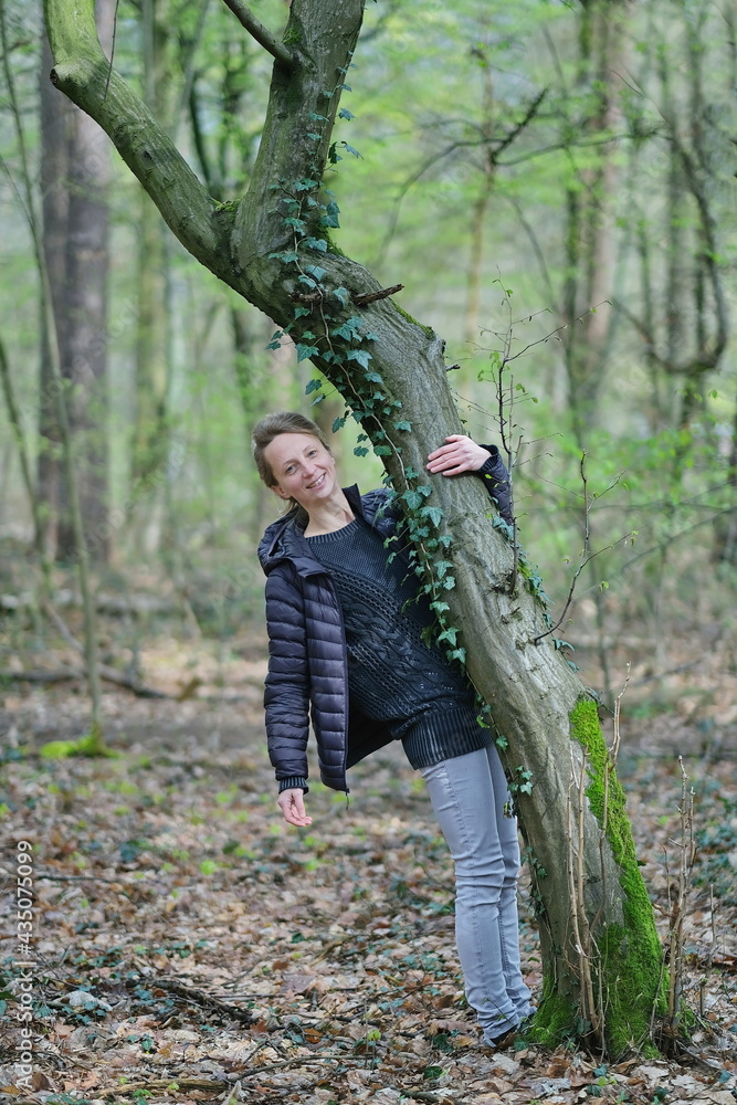 Eine junge blonde, schlanke Frau in helle jeans und dunkle blaue Jacke an einem Baum mit wenige hell grüne Blätter in Wald. Eine junge blonde, schlanke Frau in helle jeans und dunkle blaue Jacke an einem Baum mit wenige hell grüne Blätter in Wald.