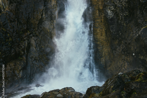 Atmospheric minimal landscape with vertical big waterfall on rock mountain wall. Powerful large waterfall in dark gorge. Nature background of high vertical turbulent falling water stream on wet rocks.