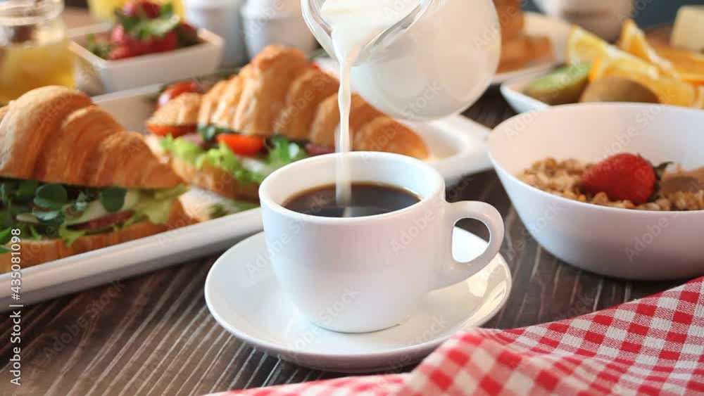 Woman hand pouring milk in to a cup of coffee. Croissant sandwiches with ham and cheese, boiled eggs, muesli and toasts on the table. Breakfast concept. Woman hand pouring milk in to a cup of coffee. Croissant sandwiches with ham and cheese, boiled eggs, muesli and toasts on the table. Breakfast concept.