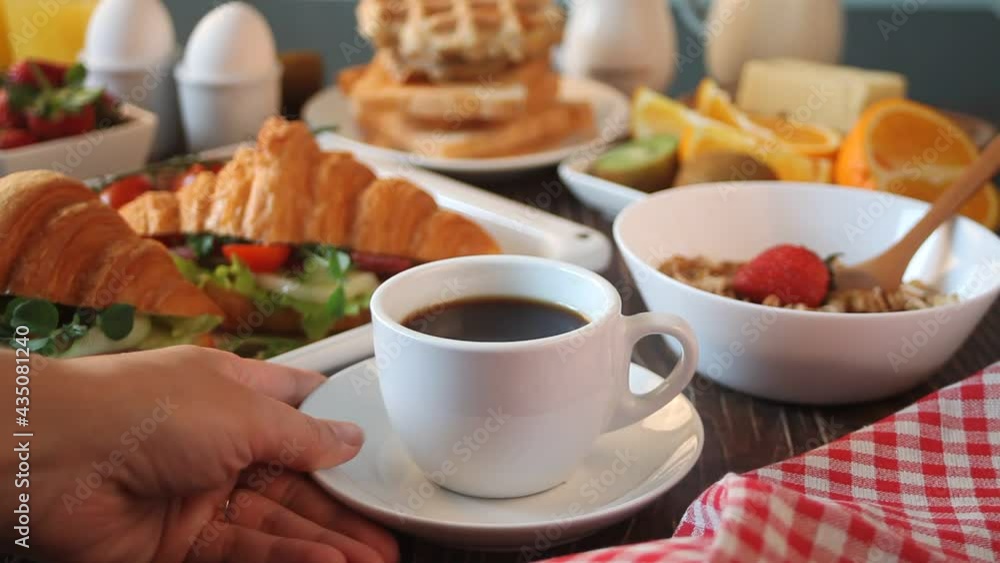 Woman hand put a cup of coffee on the table. Nearby baking sheet with croissant sandwiches with ham, cheese and greens, boiled eggs, muesli and toasts. Breakfast concept Woman hand put a cup of coffee on the table. Nearby baking sheet with croissant sandwiches with ham, cheese and greens, boiled eggs, muesli and toasts. Breakfast concept