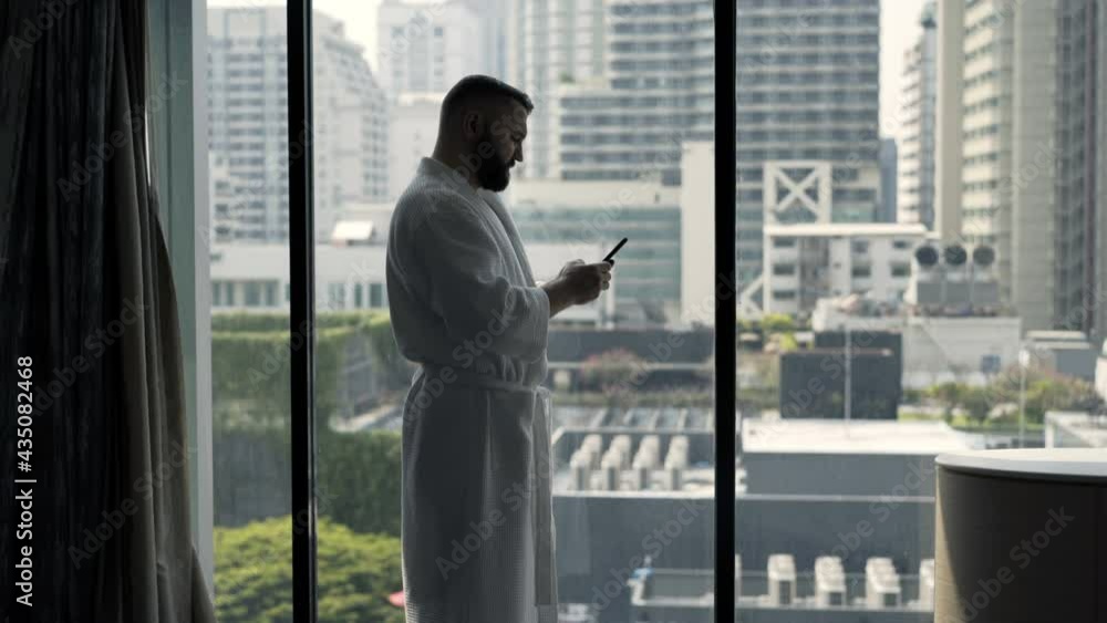 Young man in bathrobe texting on smartphone standing by window at home Young man in bathrobe texting on smartphone standing by window at home