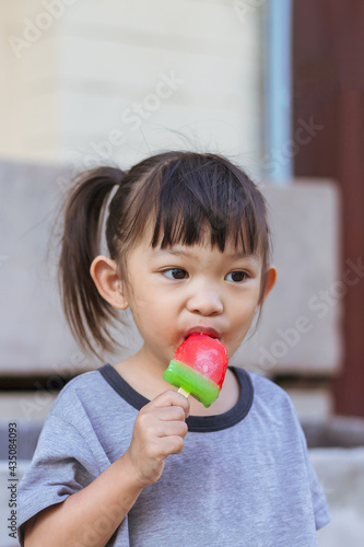 Vertical Portrait image of 3-4 years old baby. Happy Asian child girl eating and biting a red ice cream. Summer season, Delicious feeling, childhood sloppy face.