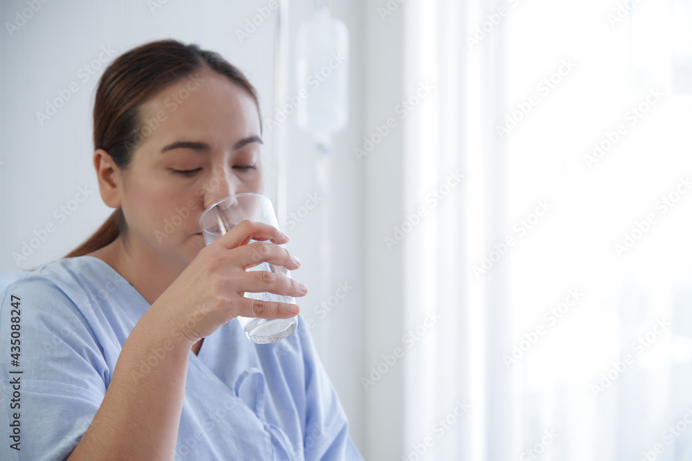 Selective focus on Asian young patient woman's hand holding a glass of water. She is drinking pure water and sitting on bed with saline bag in hospital room. Selective focus on Asian young patient woman's hand holding a glass of water. She is drinking pure water and sitting on bed with saline bag in hospital room.