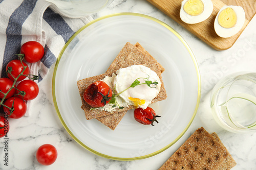 Fresh rye crispbreads with poached egg, cream cheese and grilled tomatoes on white marble table, flat lay