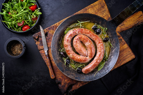 Traditional fried Italian salsiccia fresco meat sausage served with rocket salad and olives as top view in a cast iron pan photo