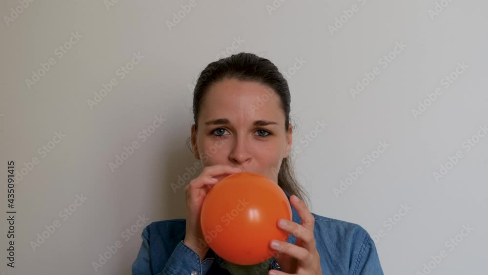 Pretty woman of Caucasian European appearance inflates orange balloon with her mouth on white background. Girl prepares for holiday and inflates balloon and then throws it forward into camera. Pretty woman of Caucasian European appearance inflates orange balloon with her mouth on white background. Girl prepares for holiday and inflates balloon and then throws it forward into camera.
