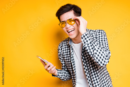 Cheerful asian guy in bright glasses holds a phone in his hand and laughs while standing on a yellow background
