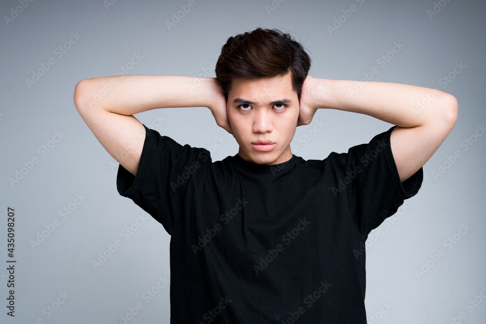 Serious Asian guy in a black T-shirt covers his ears from the noise with his hands. Isolated on light background Serious Asian guy in a black T-shirt covers his ears from the noise with his hands. Isolated on light background